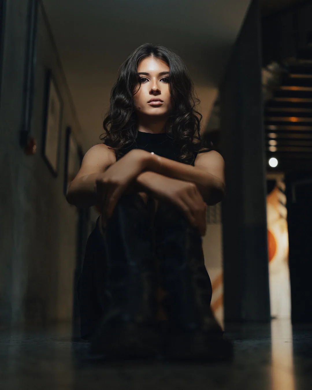 Woman with long wavy hair crouching with arms folded over her knees in a dim corridor