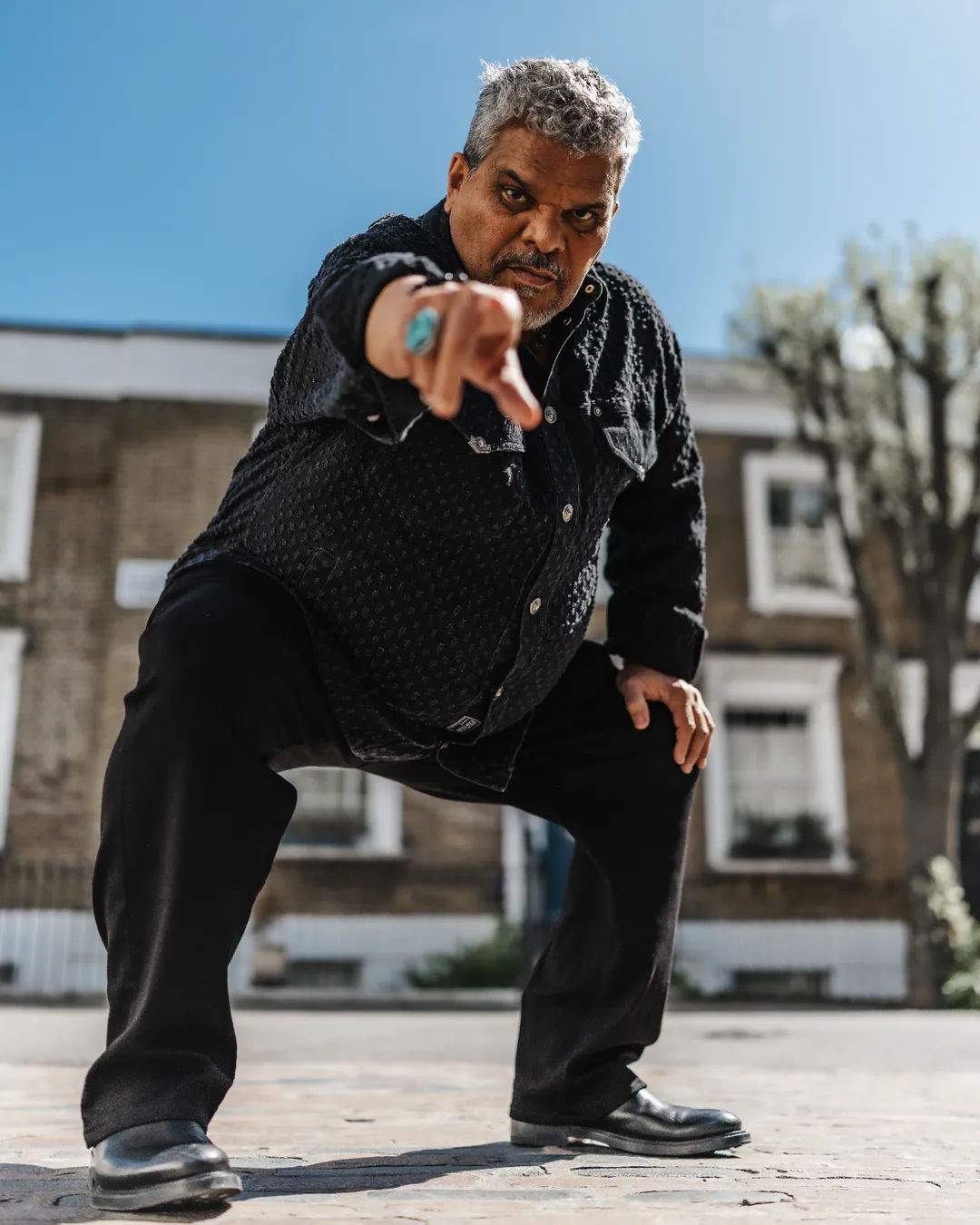 Man in a black shirt and trousers crouching low and pointing at the camera on a sunlit street