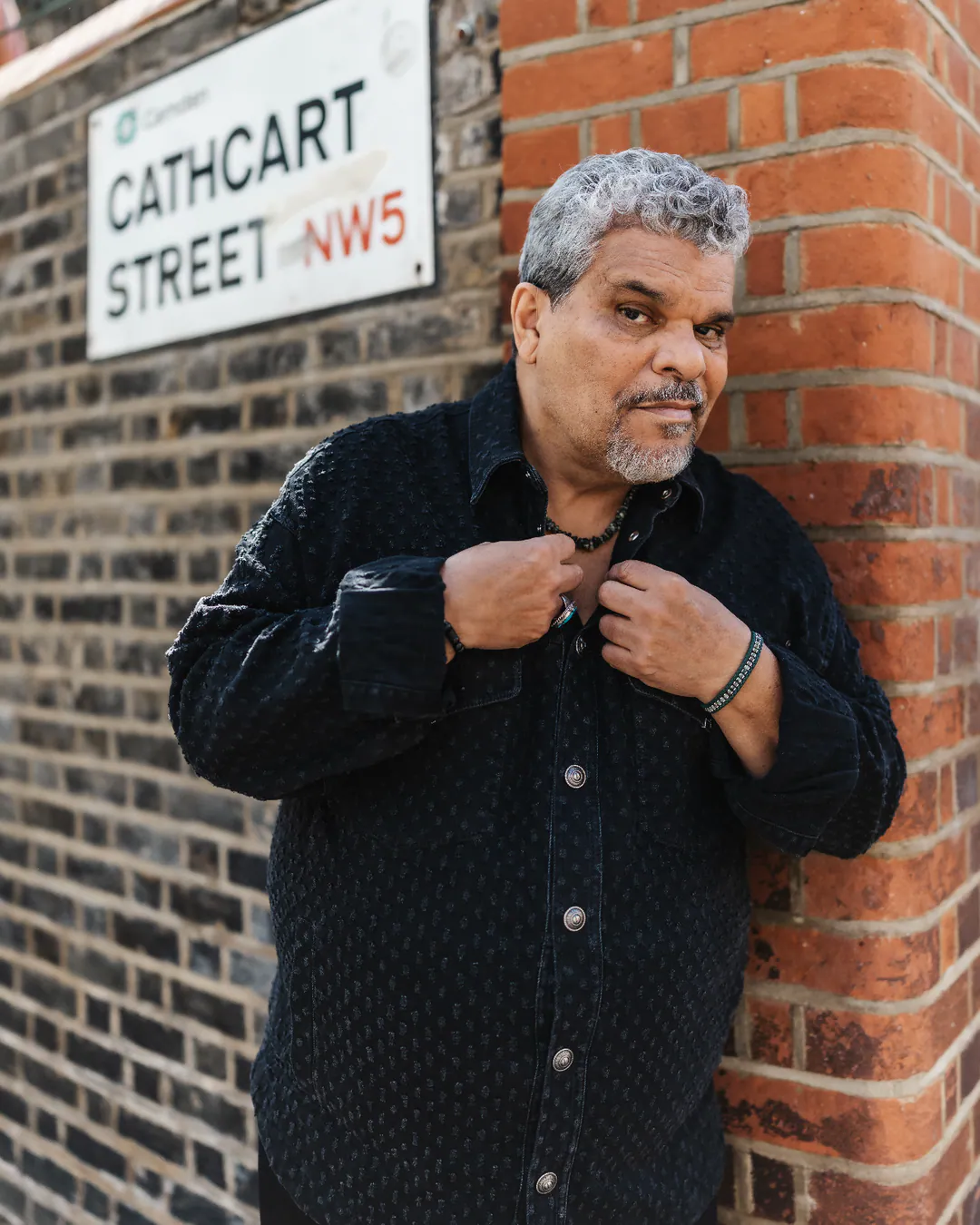 Man in a dark button-up shirt leaning against a brick wall beneath a Cathcart Street sign
