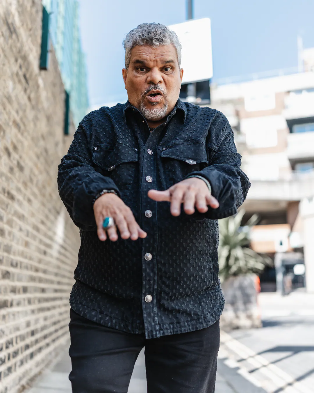 Man in a dark buttoned shirt gesturing with both hands on a sunlit street beside a brick wall