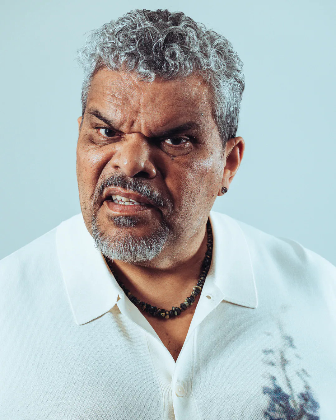Grey-haired man in a white shirt and beaded necklace scowling at the camera against a pale blue backdrop