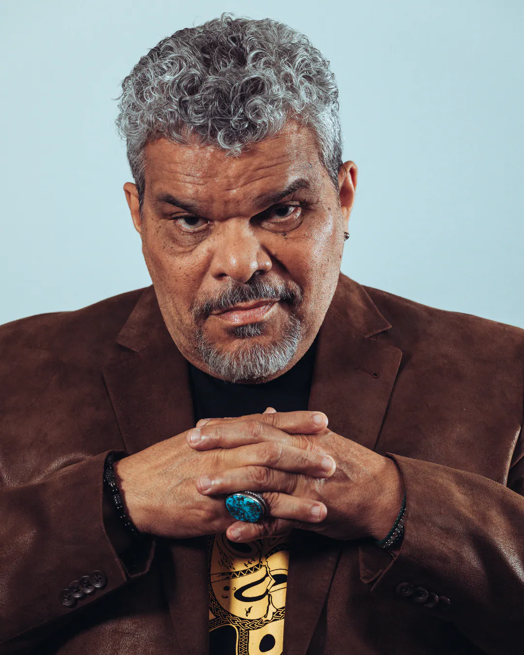 Grey-haired man in a brown blazer with hands clasped beneath his chin, serious stare, pale blue backdrop