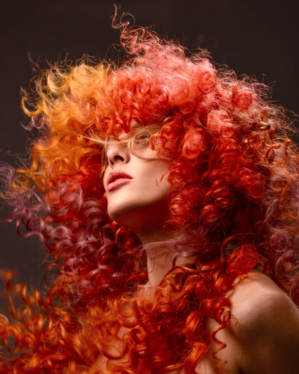 Woman with voluminous fiery red and orange curls, face tilted upwards against a dark brown backdrop