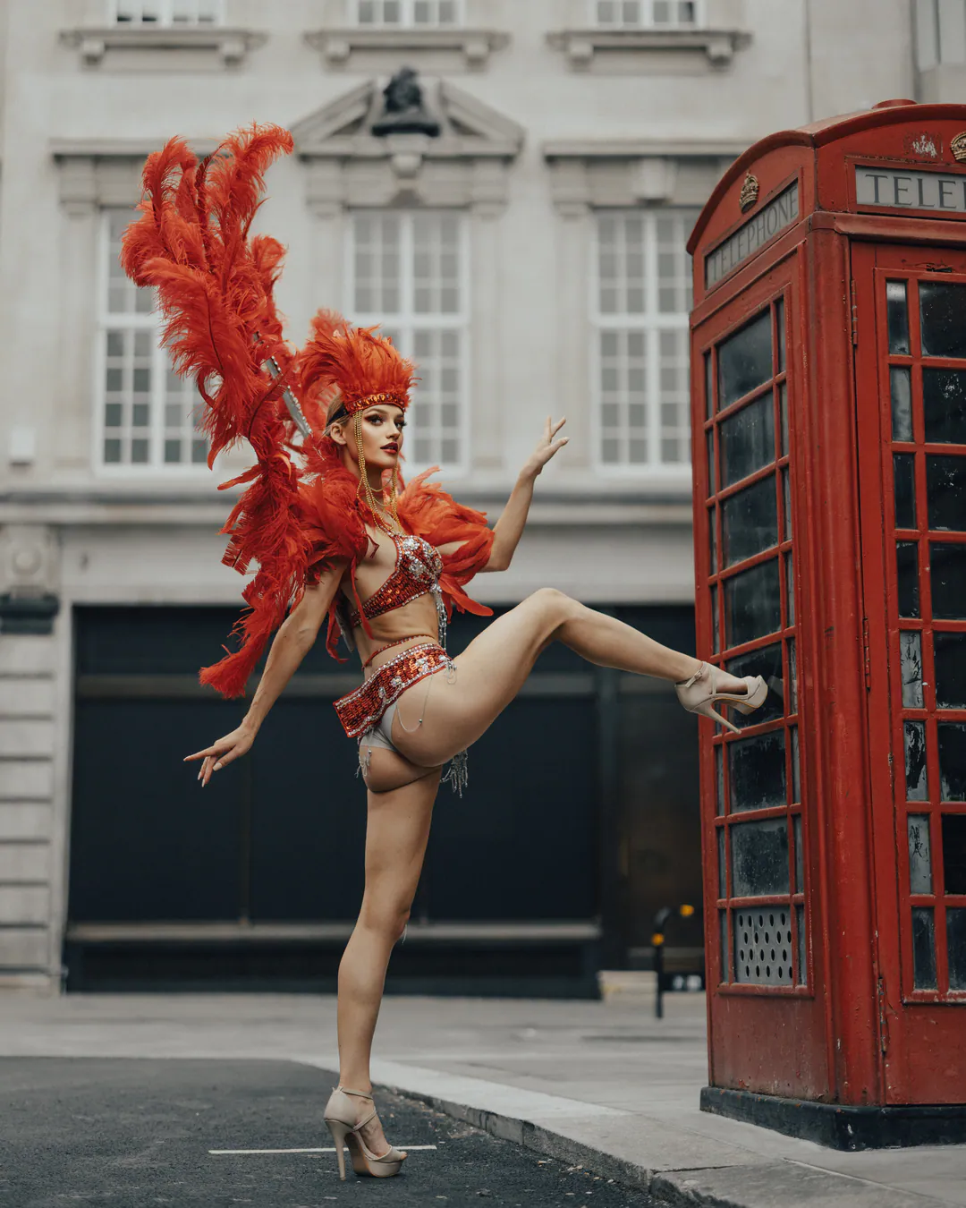 Dancer in a red feathered showgirl costume and headdress high-kicking beside a red telephone box