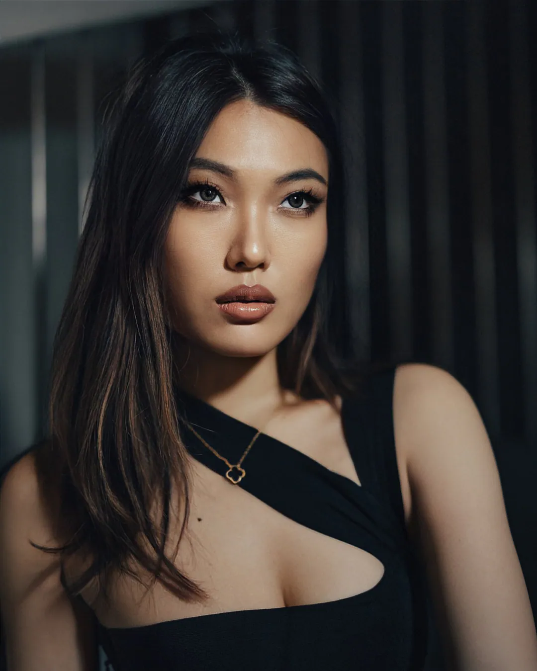 Woman with long dark hair in a black cut-out top facing the camera against a dark slatted wall