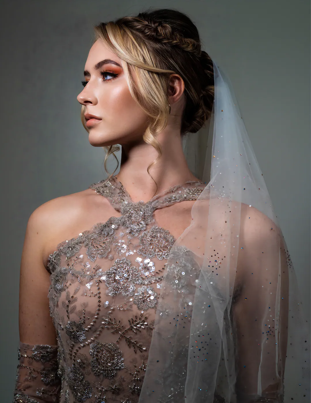 Woman in profile with a braided updo and sheer veil wearing an embellished halter gown against grey