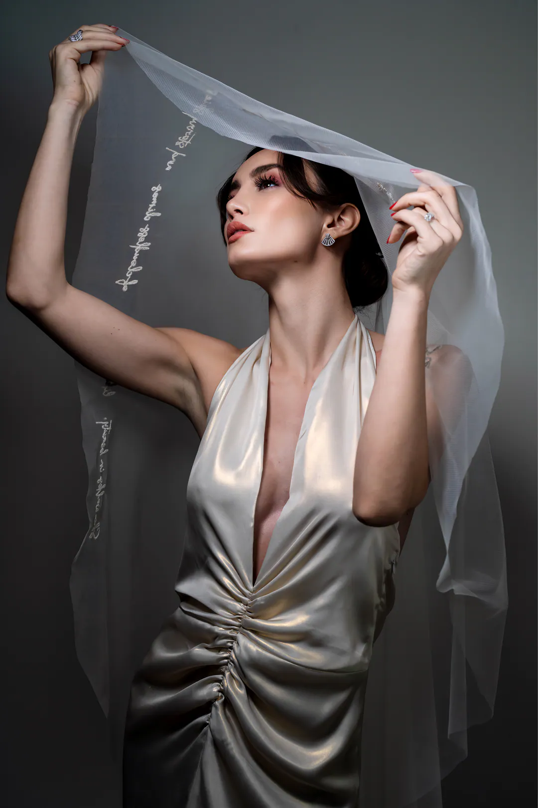 Woman in an ivory satin plunging gown holding a sheer veil above her head against a grey backdrop