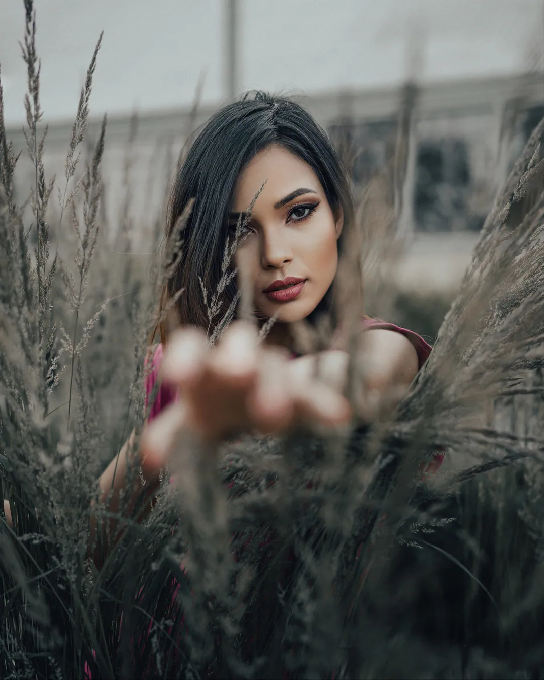Woman with long dark hair reaching a hand towards the camera through tall grasses, in muted tones
