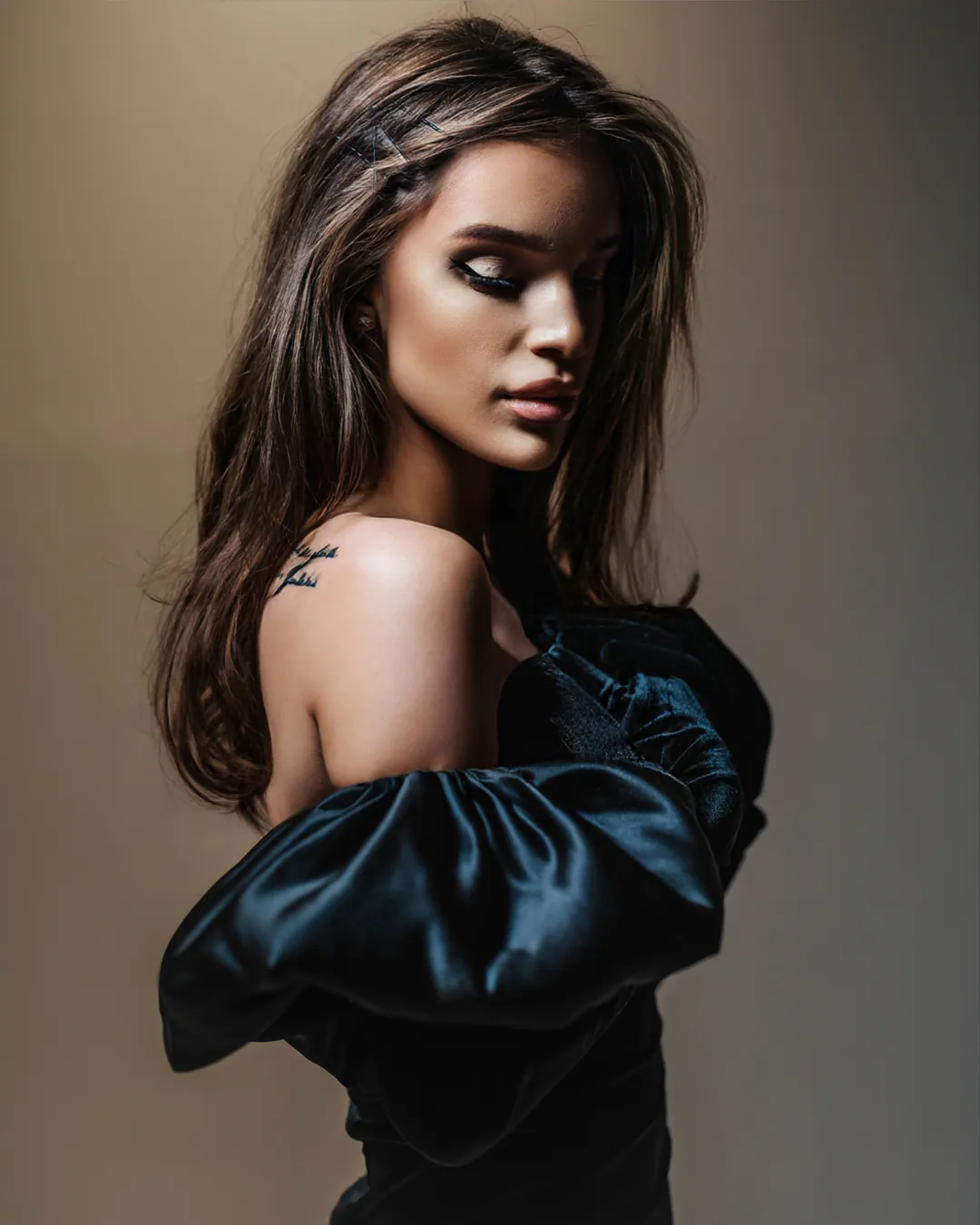 Woman in a black satin puff-sleeved dress glancing down over her bare shoulder against a beige backdrop