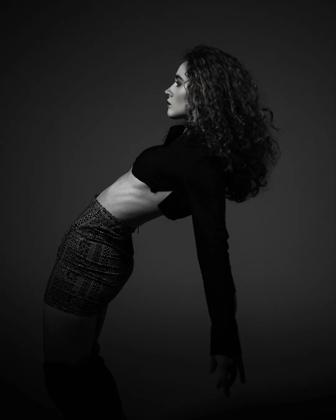 Woman with curly hair in a cropped black jacket and patterned skirt arching backwards, in black and white