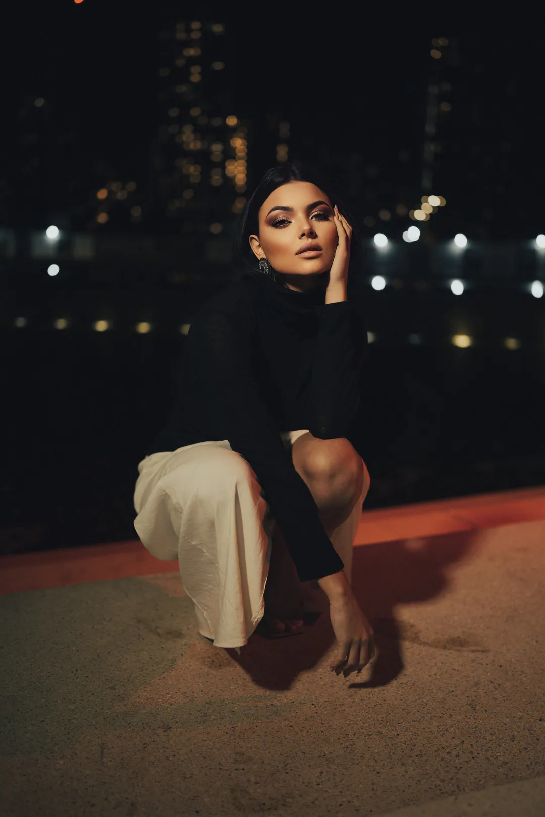 Woman in a black top and cream skirt crouching on a road at night with city lights behind