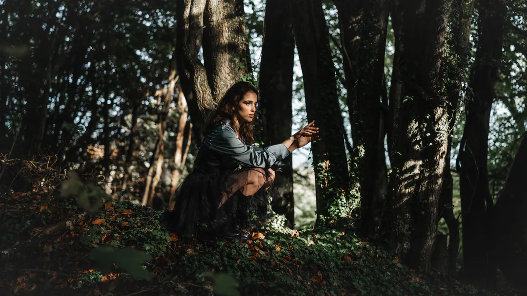 Woman crouching among tall tree trunks in a dark, shaded woodland with soft dappled light