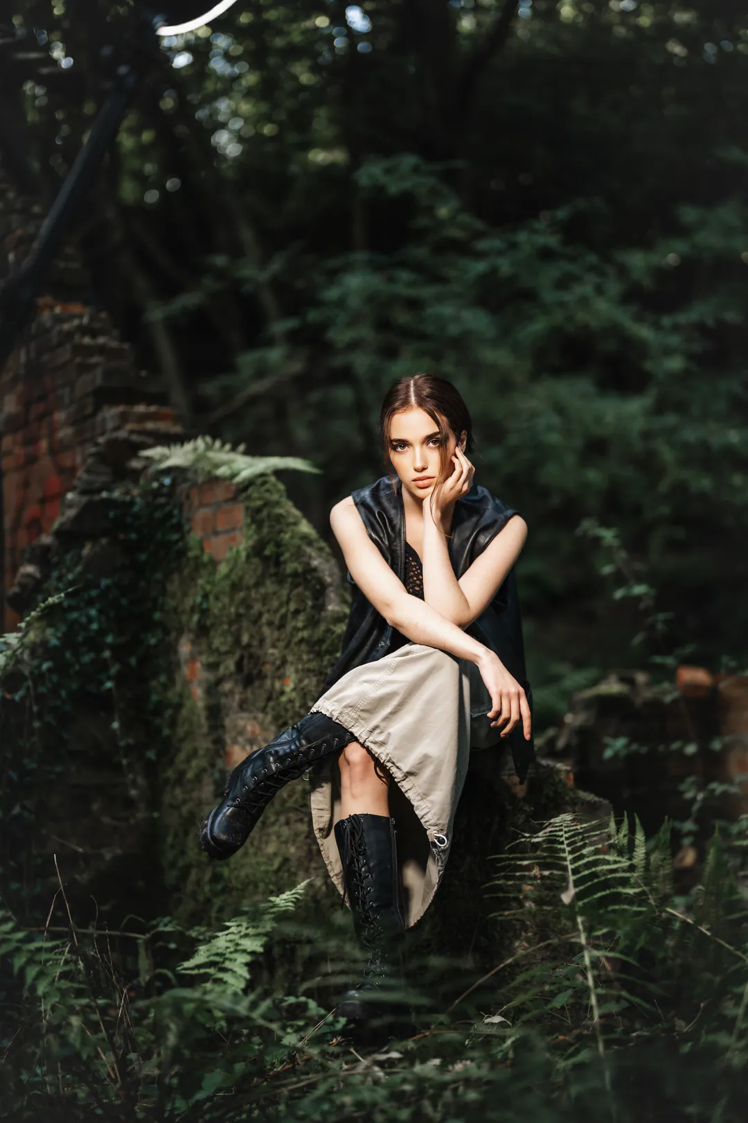 Woman in a black leather vest, pale slip skirt and black boots sitting among ferns, chin on hand