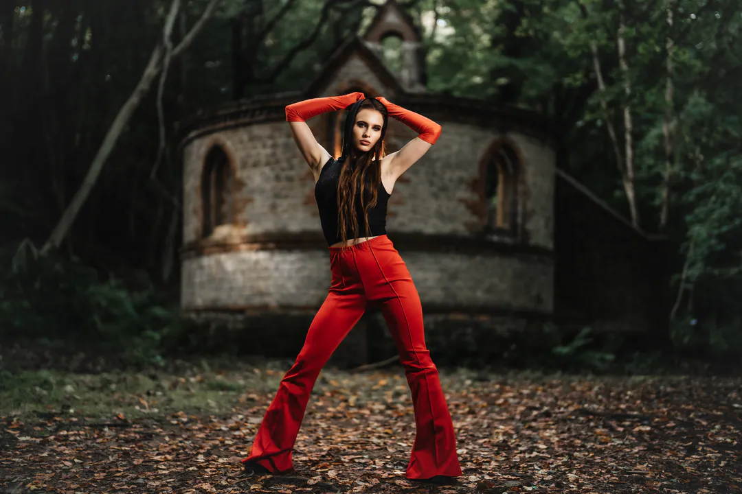 Woman in red flared trousers and red gloves striking a wide stance before a woodland chapel ruin