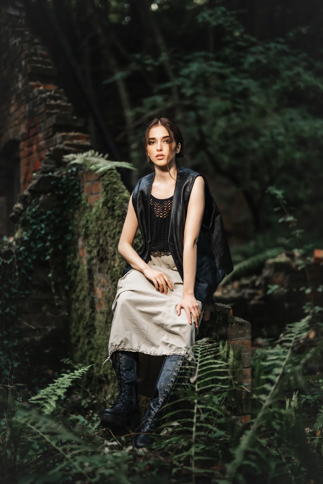Woman in a black vest and pale skirt seated on a mossy brick ruin surrounded by ferns