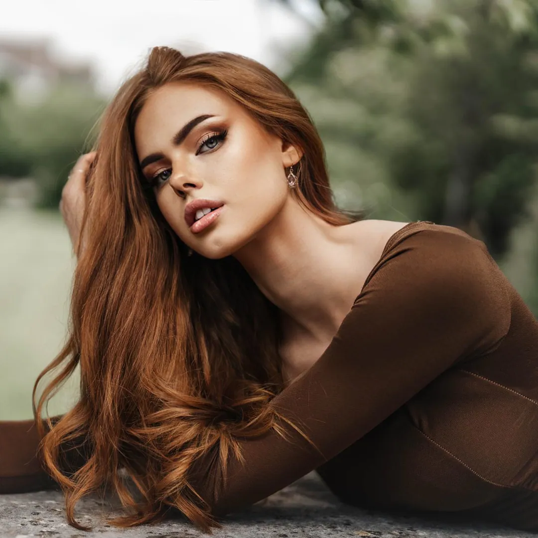 Woman with long auburn hair in a brown long-sleeved top leaning forward on a stone ledge, greenery behind