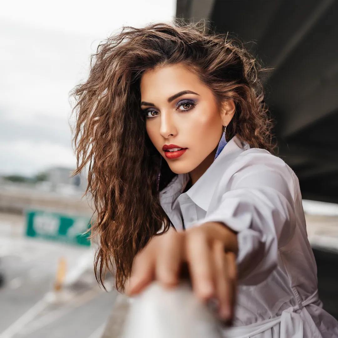 Woman with long curly hair in a white shirt reaching towards the camera from a bridge railing