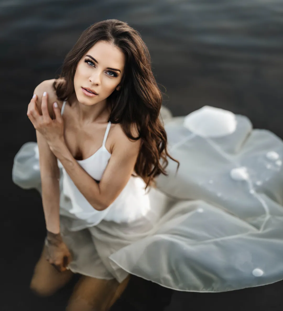 Woman in a white camisole crouching in dark water, sheer fabric floating around her, gazing at the camera