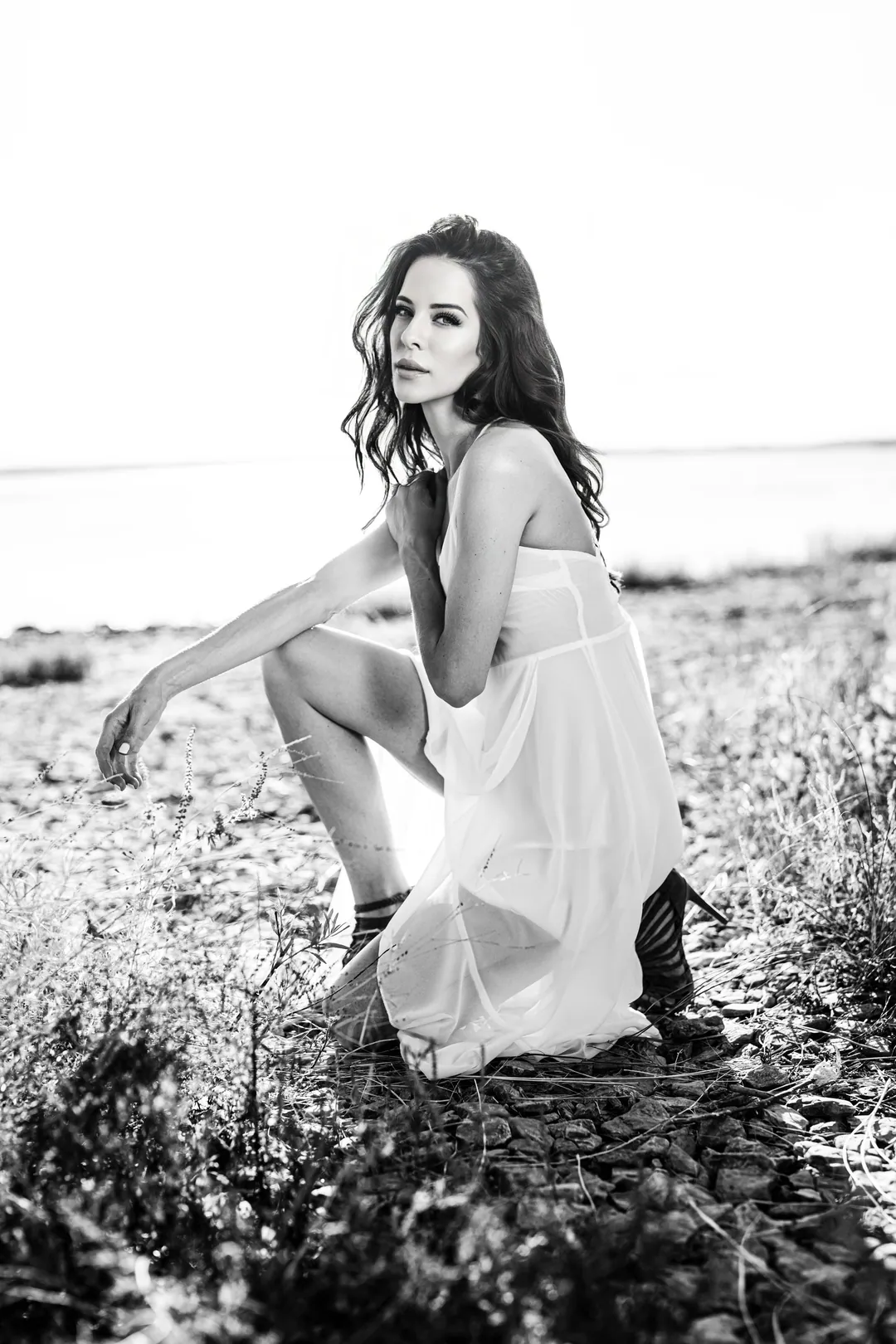 Woman in a sheer white slip dress and heeled boots crouching among coastal grasses, in black and white