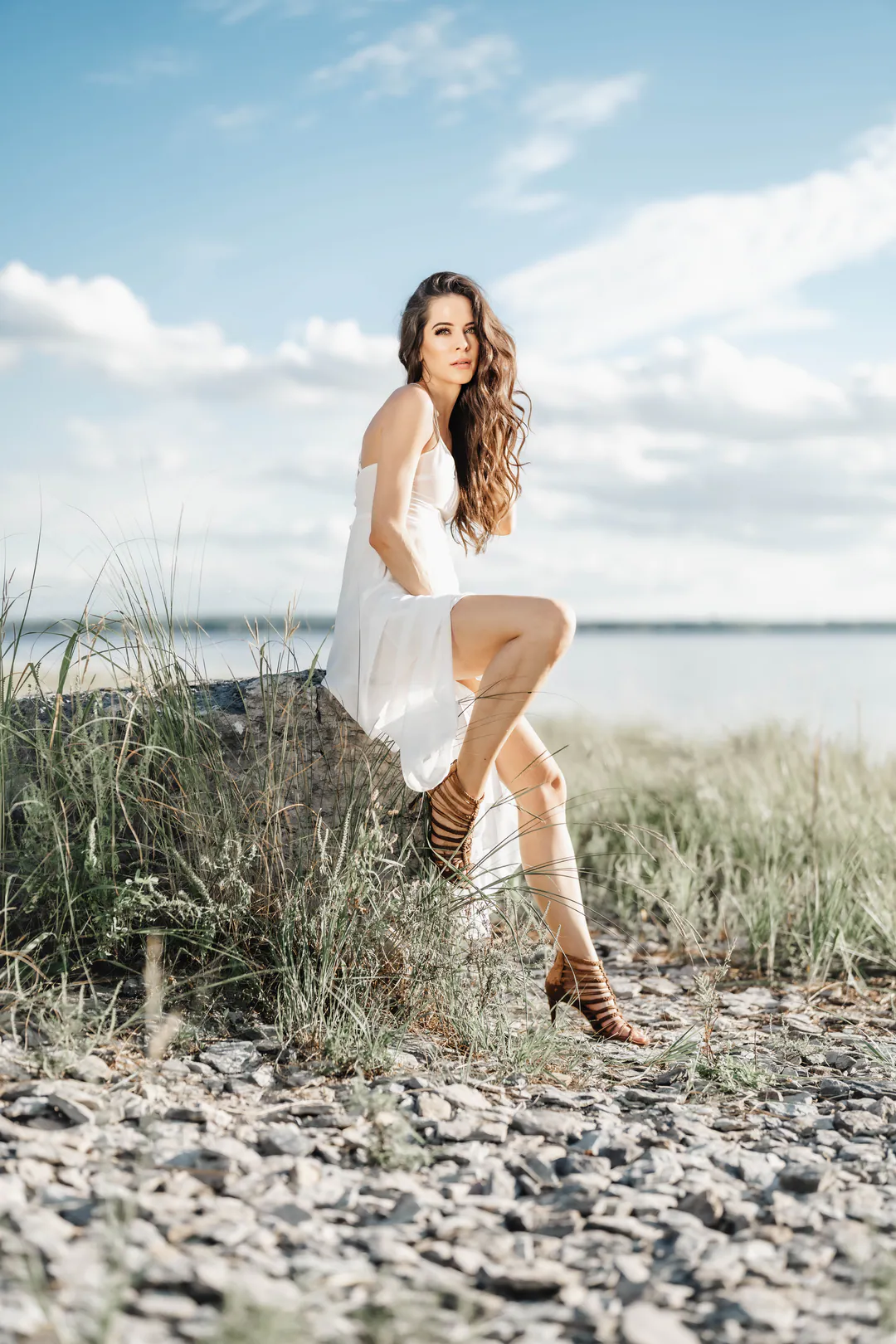 Woman in a short white dress perched on a rock among beach grasses under a bright blue sky