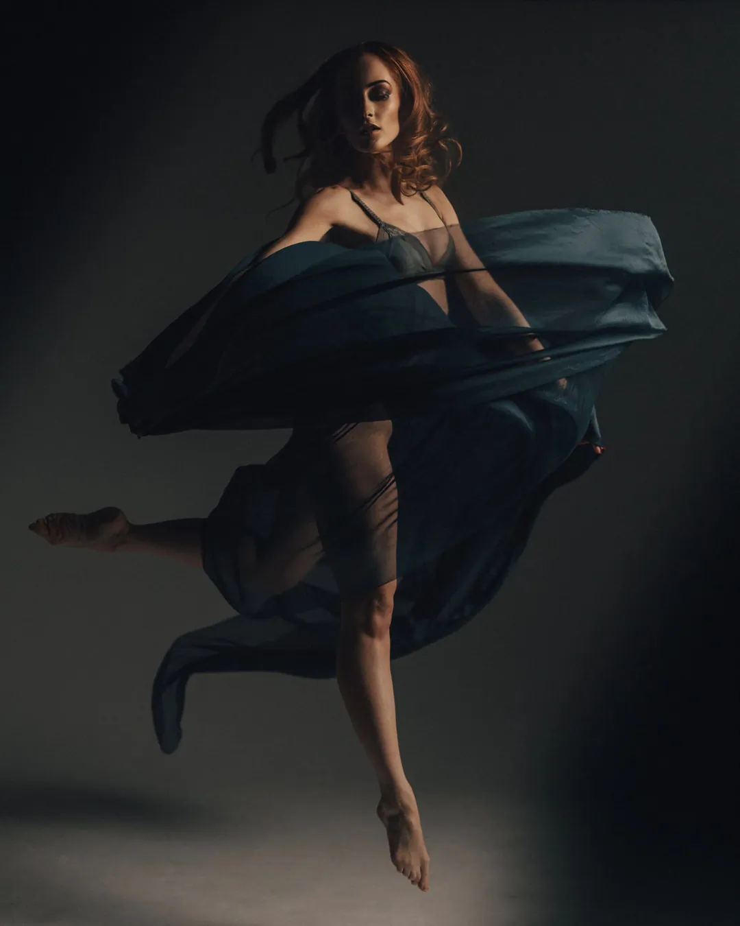 Dancer with red curls leaping barefoot as a sheet of dark blue silk swirls around her body, on a dark studio floor