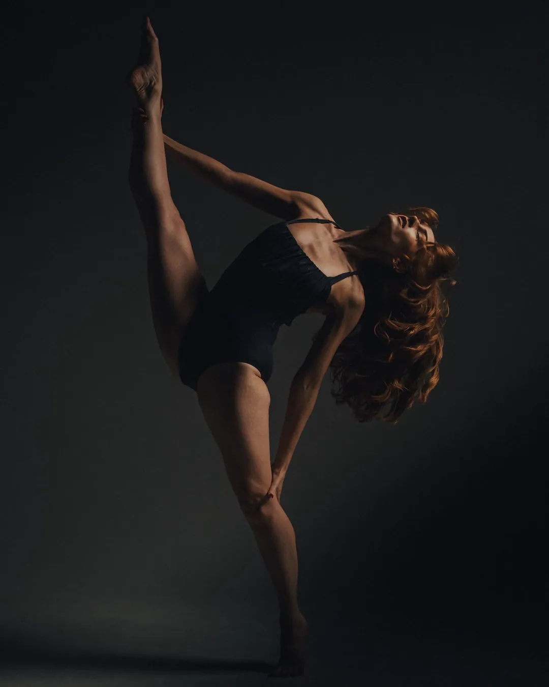Dancer in a black leotard holding a standing split, one leg raised straight overhead and red hair falling back, against shadow