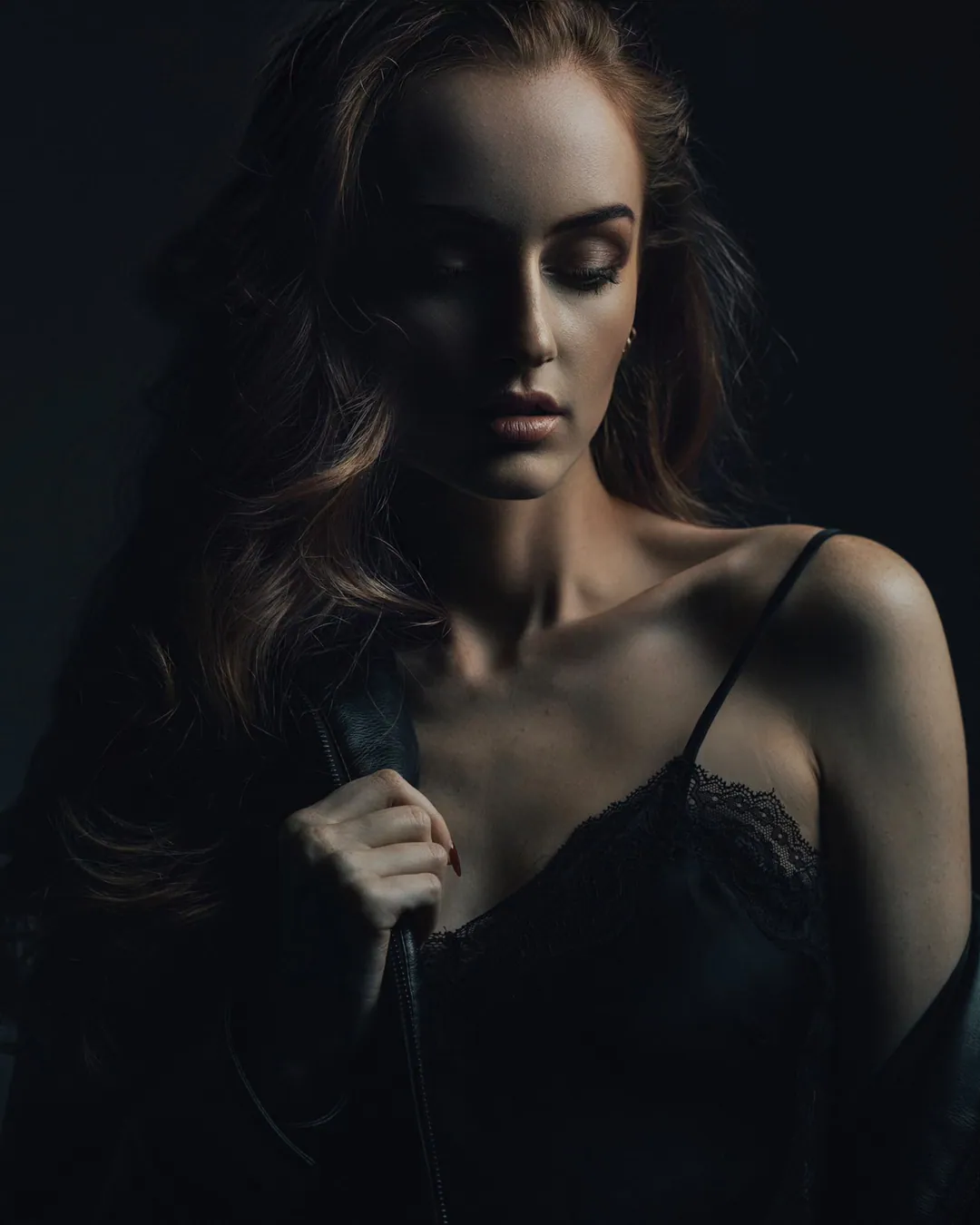 Woman with long auburn hair and eyes lowered in a black lace camisole, holding a leather jacket at her shoulder, lit from one side