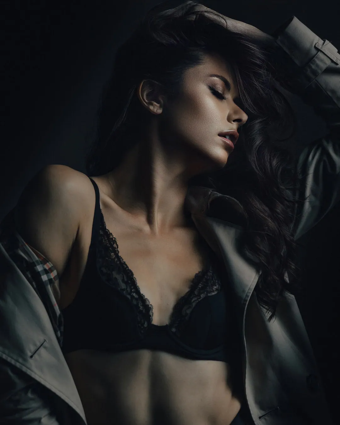 Woman with long dark hair and eyes closed, arm raised into her hair, wearing a black lace bra under an open trench coat