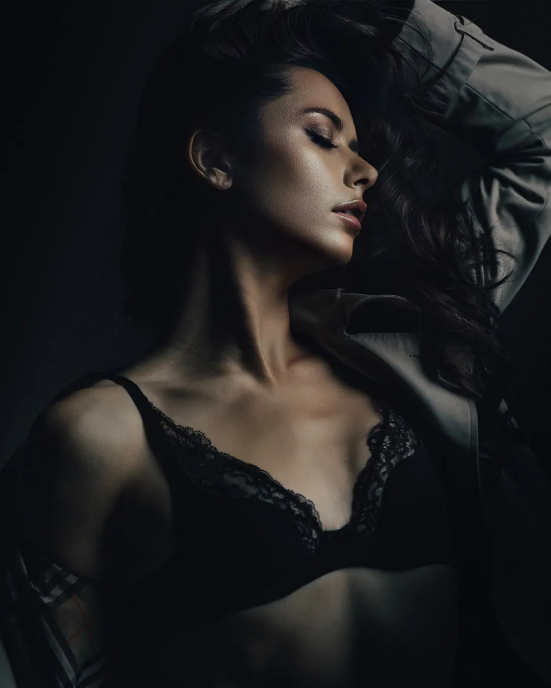 Close crop of a woman with eyes closed and head tilted back, a trench coat slipping from her shoulder over black lace