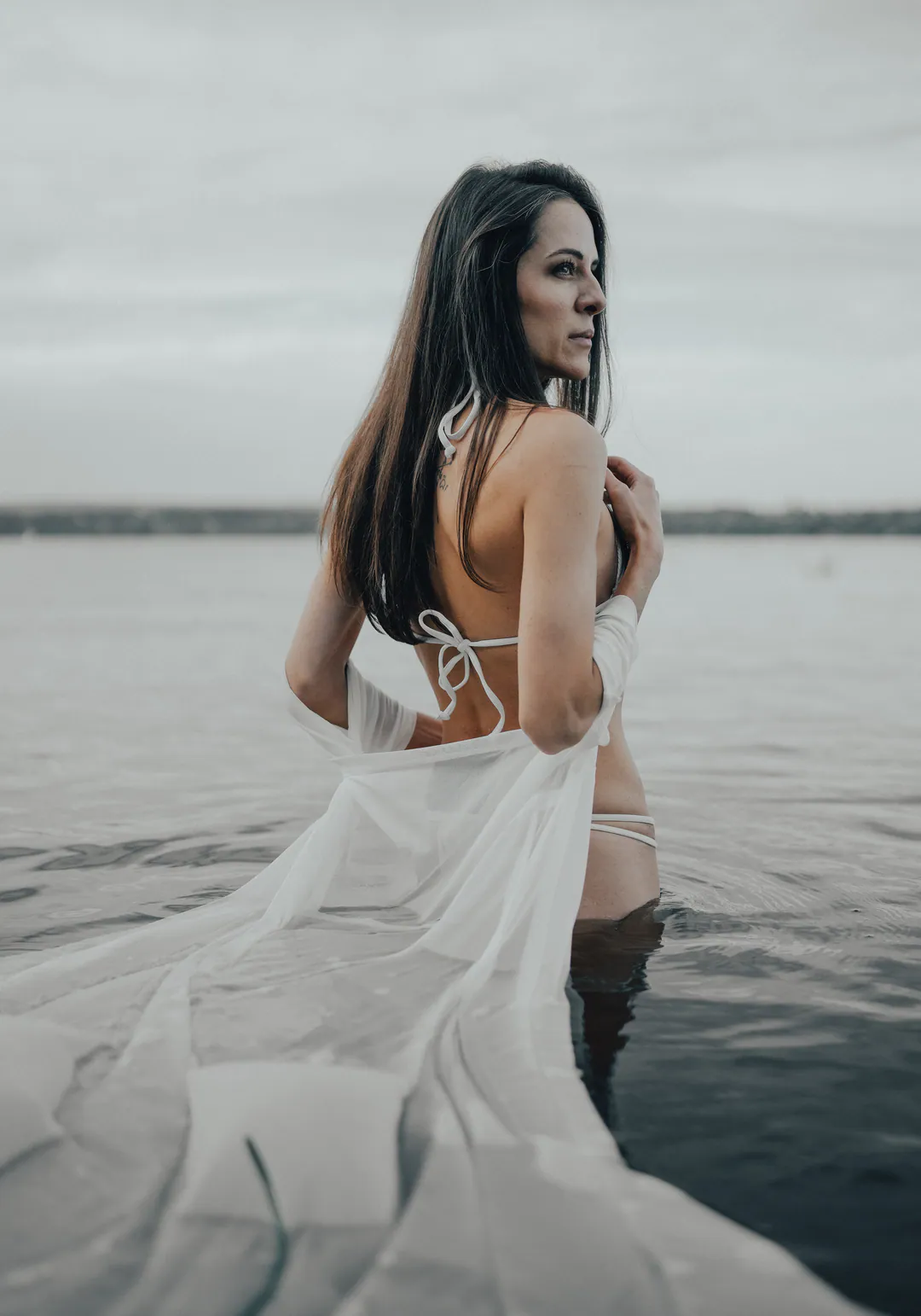 Woman in a white bikini and sheer white wrap standing waist-deep in grey water, glancing over her shoulder