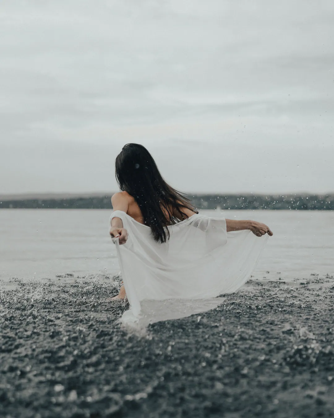 Woman in a flowing white dress stepping into shallow water, seen from behind under an overcast sky