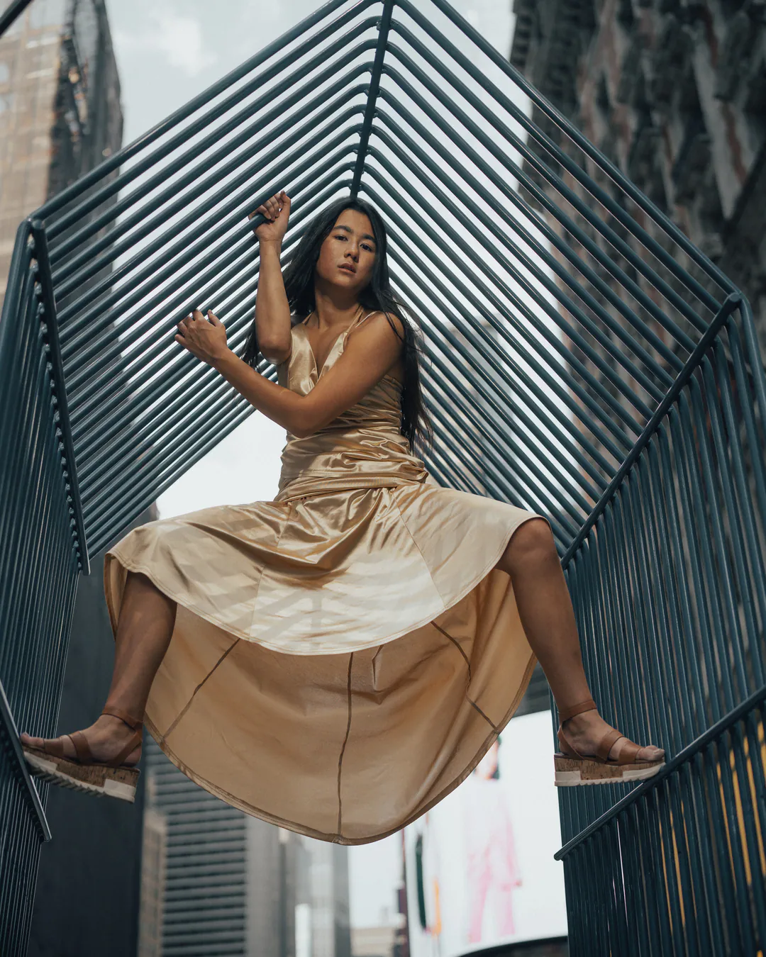 Woman in a gold satin dress perched on a slatted metal sculpture, shot from below among skyscrapers