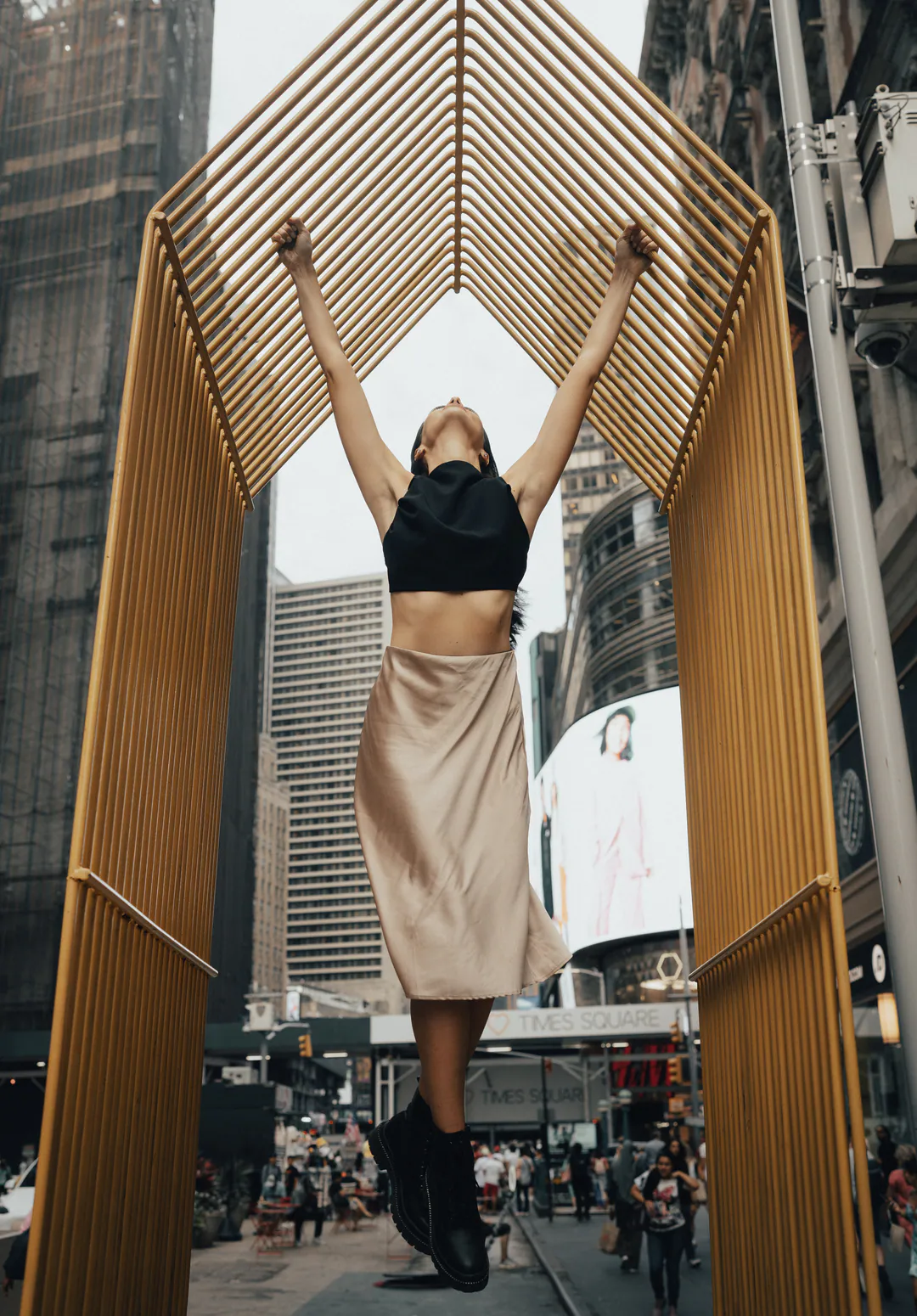 Woman in a crop top hanging from a golden arch, head tipped back, above a busy city street