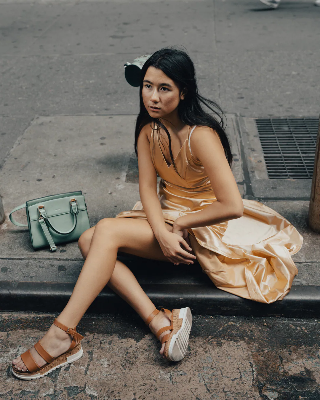 Woman in a gold satin dress and sandals sitting on a kerb beside a mint green handbag