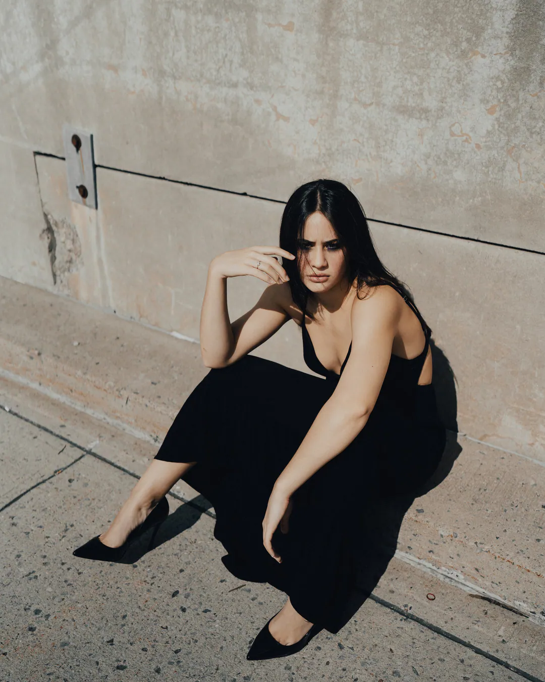 Woman in a black slip dress and pointed heels crouching against a sunlit concrete wall