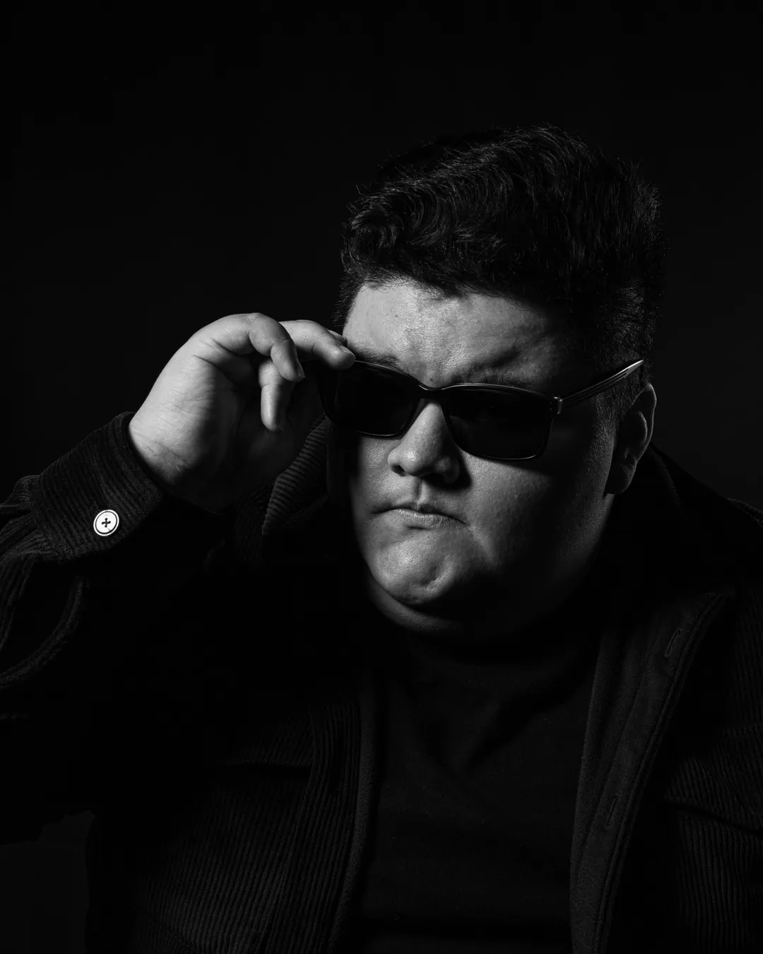 Man with a dark quiff adjusting black sunglasses with pursed lips against a dark backdrop, black and white