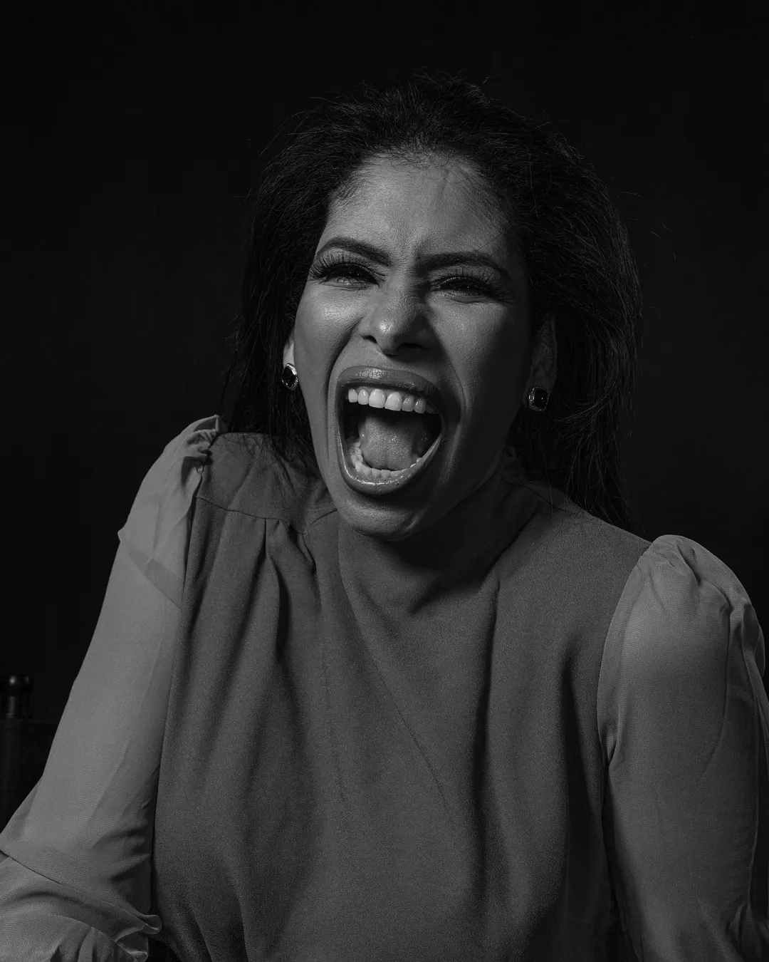 Woman in a puff-sleeved top laughing loudly with her head tilted against a dark backdrop, black and white