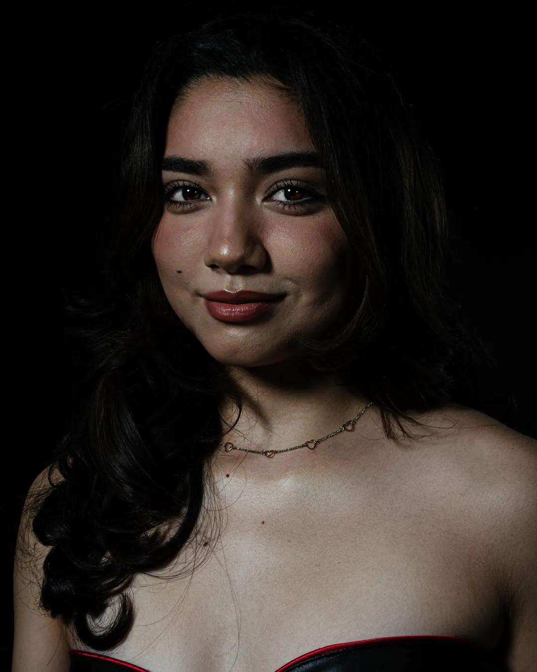 Woman with long dark wavy hair and a fine necklace smiling softly in a strapless top against black