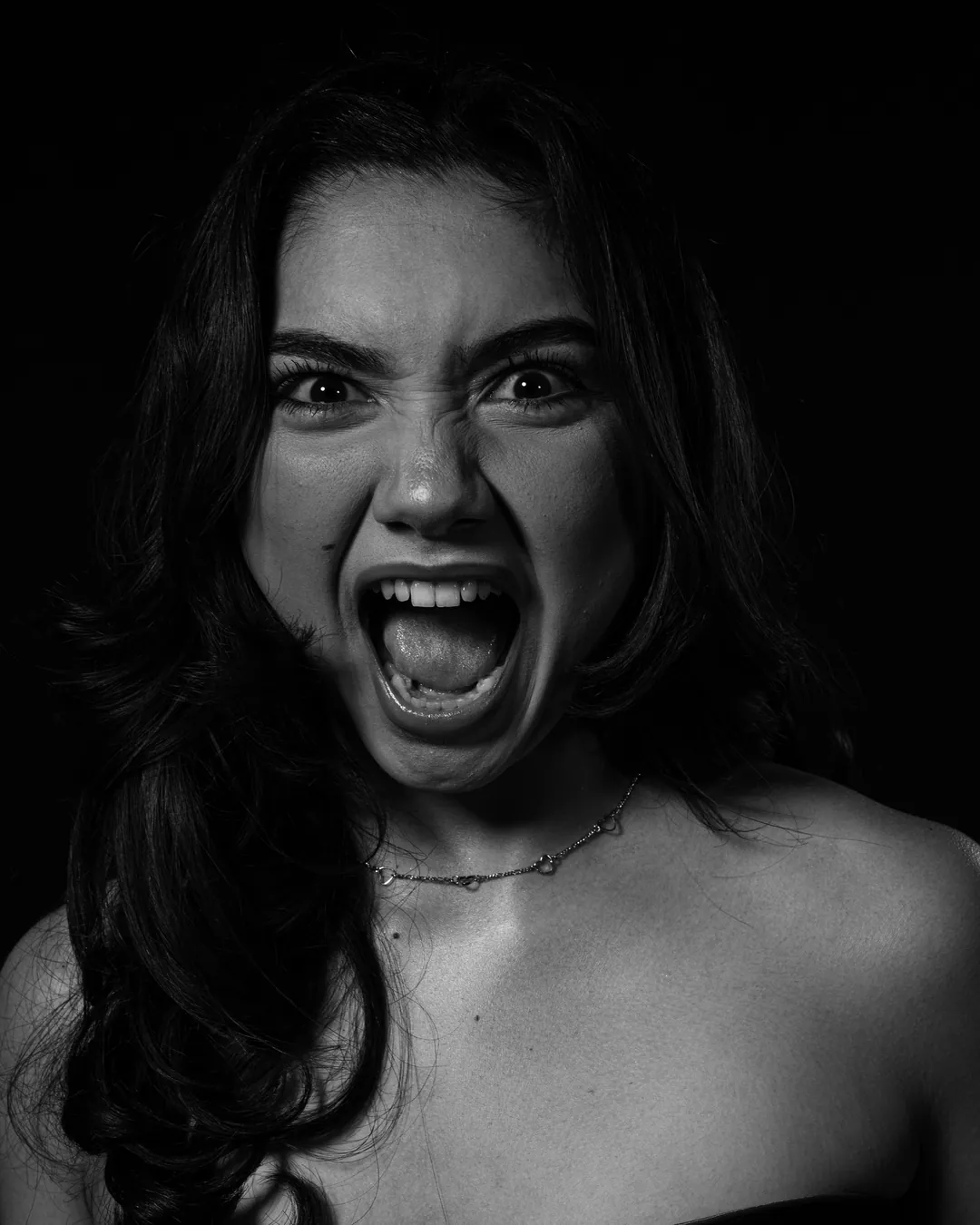 Woman with long dark hair and bare shoulders screaming towards the camera, black and white low-key portrait