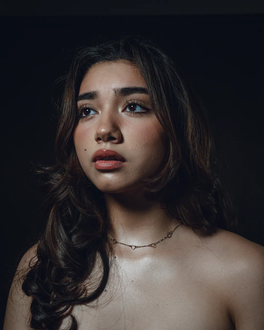 Woman with long wavy hair and bare shoulders gazing upwards in soft warm light against a dark backdrop