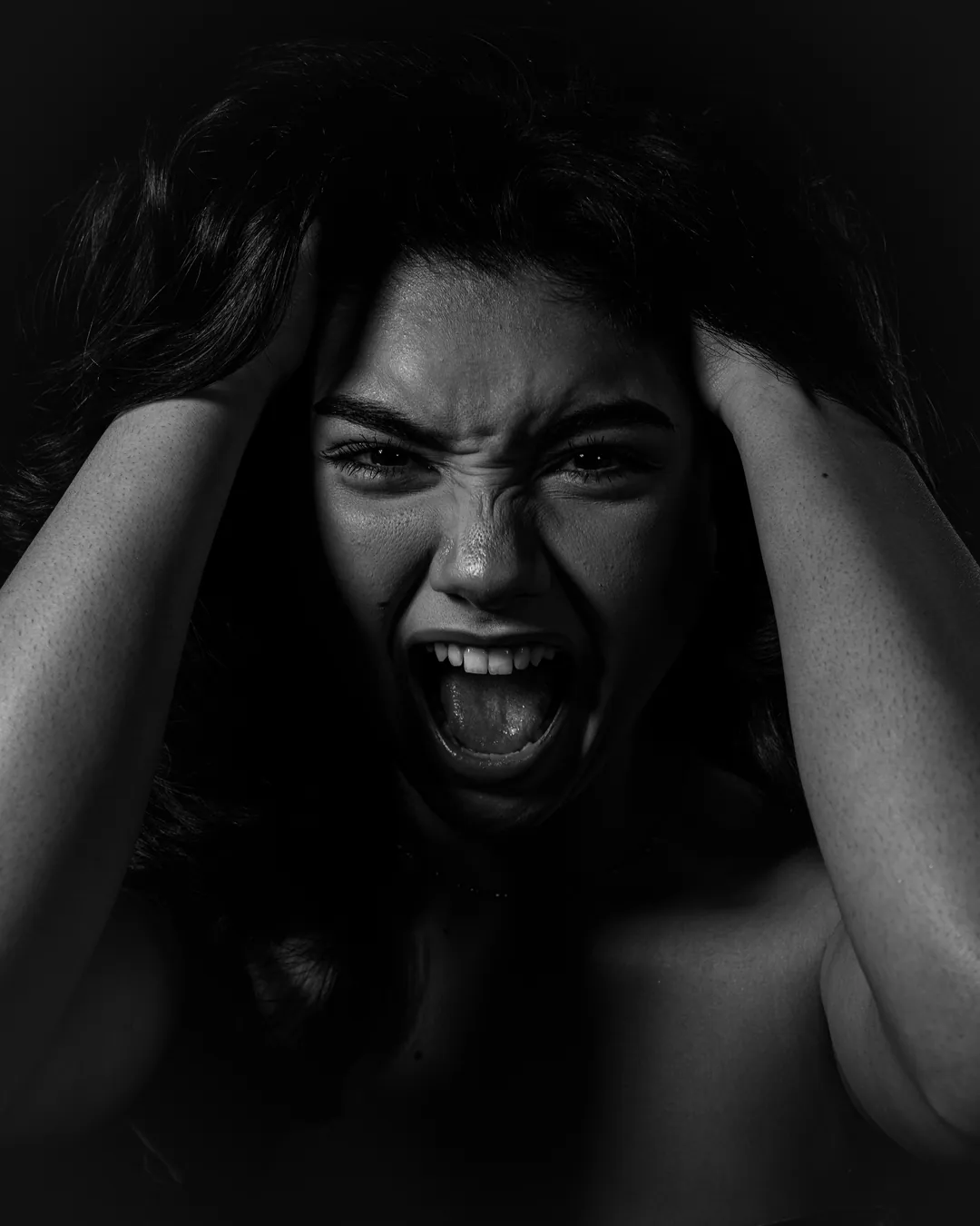 Woman gripping her hair with both hands and shouting in harsh low-key light, black and white