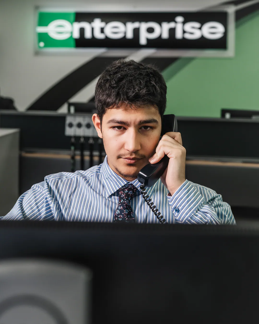 Man in a striped shirt and tie talking on a desk phone beneath an Enterprise sign