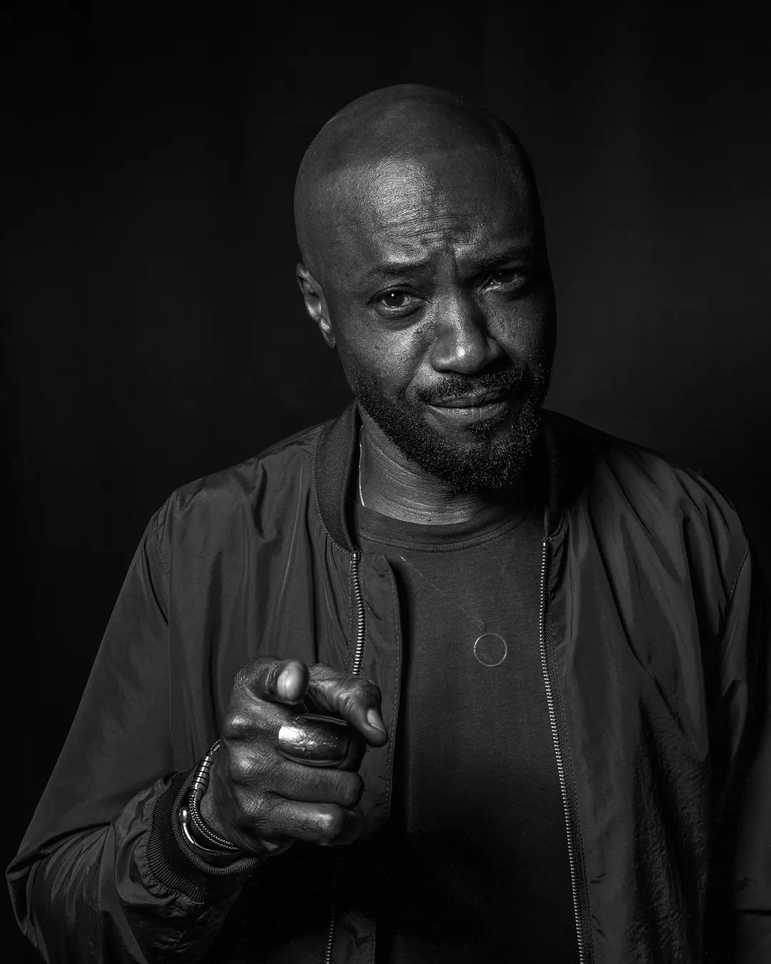 Bearded man in a bomber jacket pointing at the camera with a wry smile, black and white