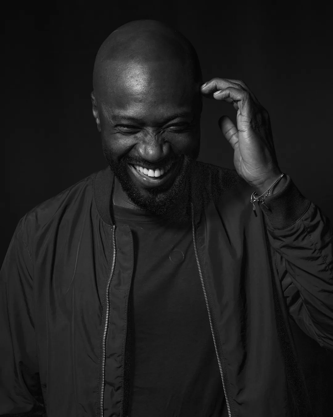 Bearded man in a bomber jacket grinning and scratching his head against a dark backdrop, black and white