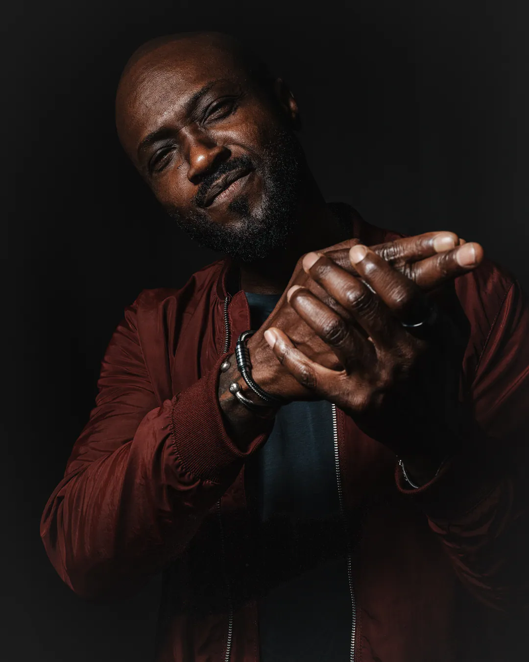 Man in a red bomber jacket making finger guns with his head tilted, moody low-key light on black