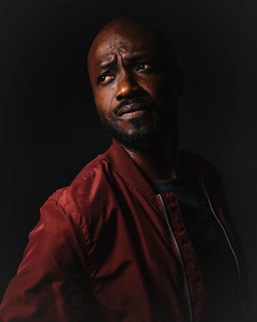 Man in a red bomber jacket glancing sideways with a sceptical look, dramatic low-key light on black