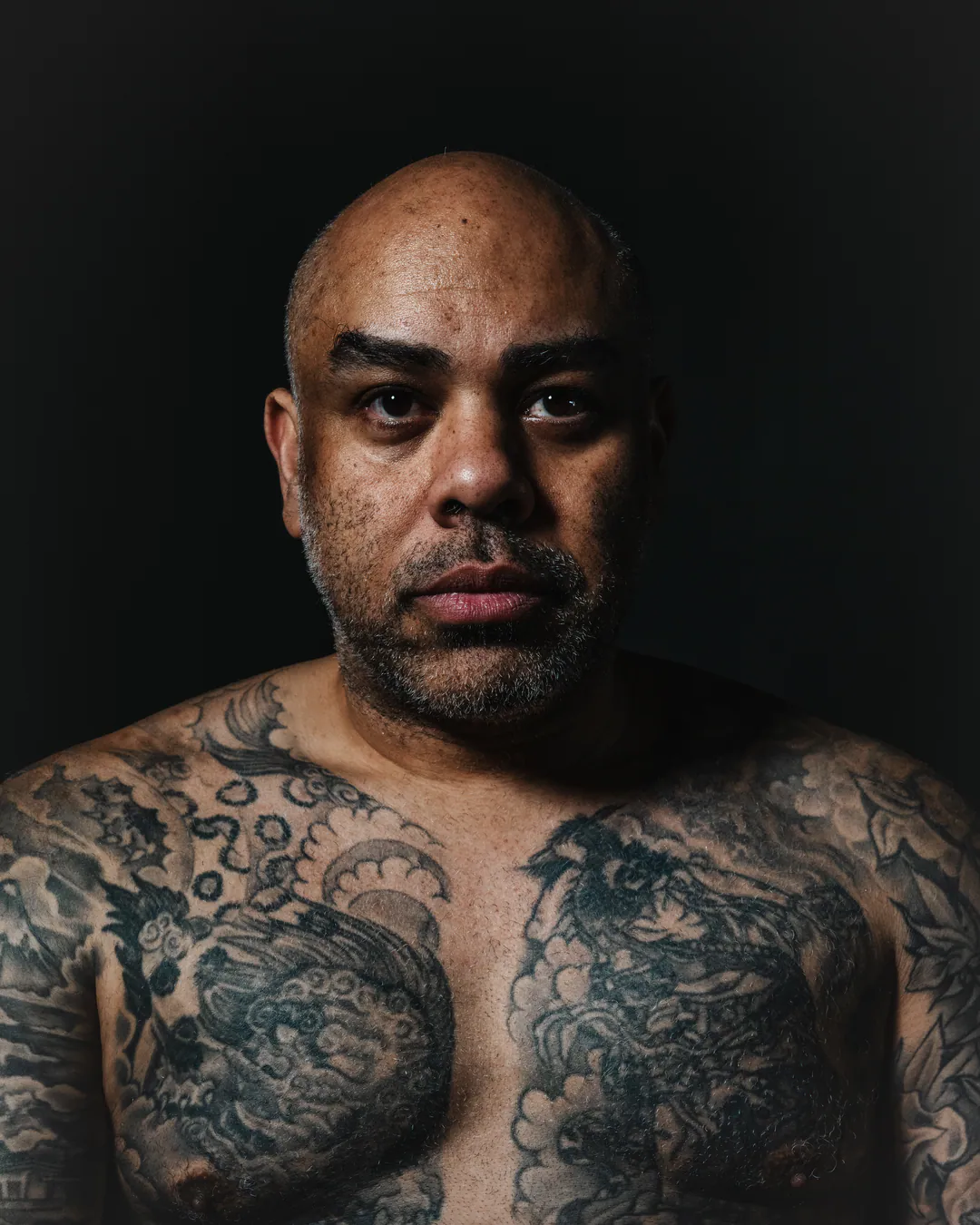 Bald man with a tattooed chest and shoulders looking straight at the camera against a dark backdrop