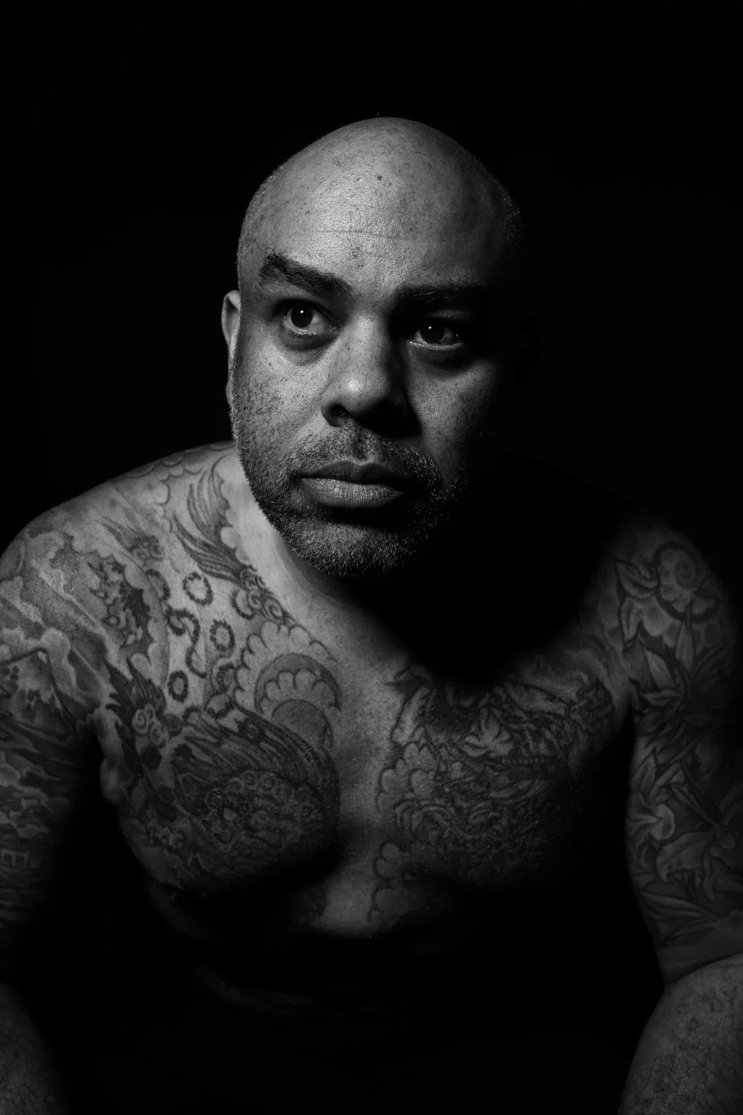 Bald man with a heavily tattooed chest leaning forward with an intense stare, black and white