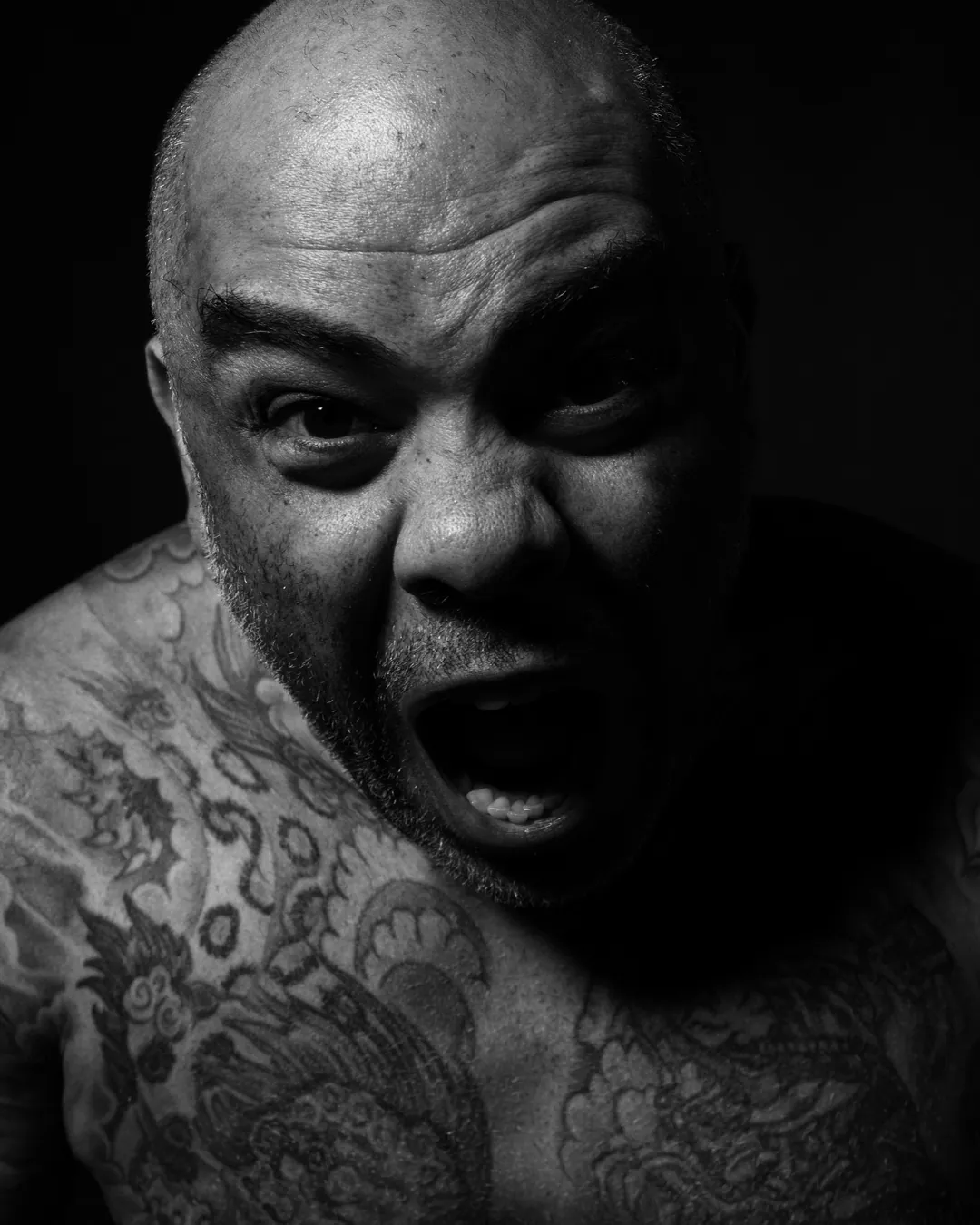Bald, tattooed man shouting towards the camera in a tight close-up with hard side light, black and white