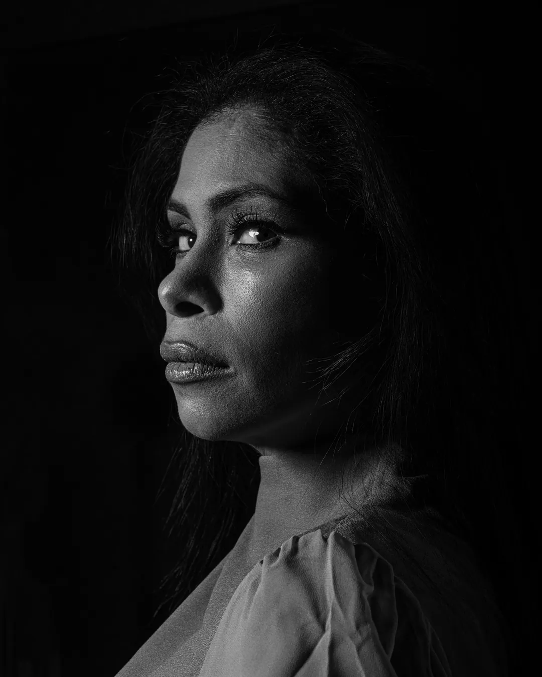 Woman in near profile glancing towards the camera, side-lit against a deep black backdrop, black and white